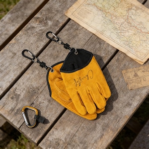 May include: Mustard yellow work gloves with black trim and the name "Christoff" in cursive script. The gloves are attached to a black carabiner clip. A gold and silver carabiner clip is also visible.