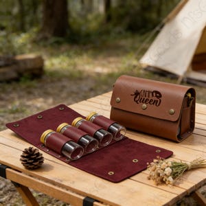 May include: A brown leather spice rack with four glass bottles, gold lids, and burgundy leather sleeves. The rack is open, revealing the bottles, and sits on a wooden table next to a matching leather bag with the text "Camp Queen".