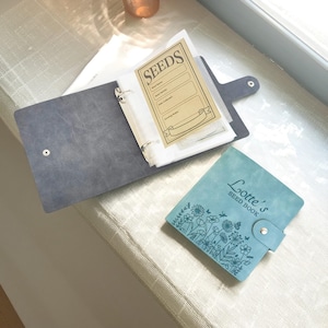 May include: Open, gray seed binder with a button closure, holding seed packets. A smaller, teal seed book with the text "Lotte's Seed Book" and floral illustrations is next to it. The binder is on a light-colored surface.