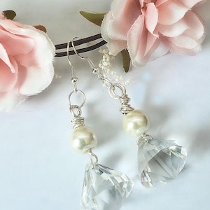 May include: Elegant dangle earrings featuring a faux pearl and a faceted crystal pendant. The earrings are silver-toned and have a delicate wire-wrapped design. The background includes soft pink flowers.