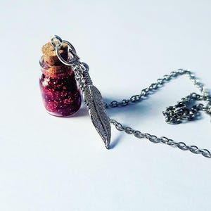 May include: A silver necklace featuring a small glass bottle filled with red glitter and a cork stopper. A silver feather charm hangs from the chain. The necklace is displayed on a white surface.