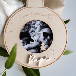 Pu&ograve; includere: Un ornamento natalizio in legno a forma di sfera con una cornice per foto al centro. L'ornamento &egrave; decorato con un piccolo albero e le parole "Frohe Weihnachten" e "Papi".