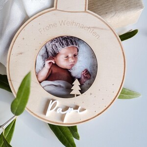 Pu&ograve; includere: Un ornamento natalizio in legno a forma di sfera con una cornice per foto al centro. La cornice contiene una foto di un bambino con un berretto di lana. L'ornamento &egrave; inciso con il testo "Frohe Weihnachten" e "Papi".