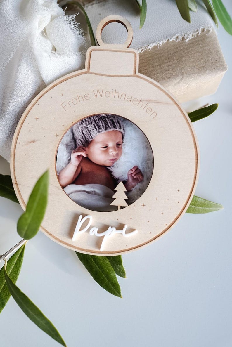 Pu&ograve; includere: Un ornamento natalizio in legno a forma di sfera con una cornice per foto al centro. La cornice contiene una foto di un bambino con un berretto di lana. L'ornamento &egrave; inciso con il testo "Frohe Weihnachten" e "Papi".