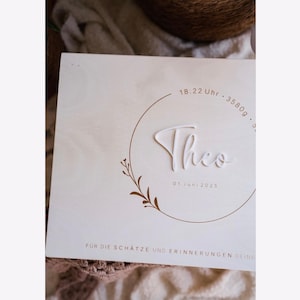Könnte beinhalten: Eine hellfarbene Holzkiste mit dem Namen "Theo" in 3D-Schrift. Die Kiste hat ein kreisförmiges Design mit Uhrzeit, Gewicht und Datum. Ein florales Design befindet sich unten links. Der Text "FUR DIE SCHÄTZE UND ERINNERUNGEN DEINE" steht unten.