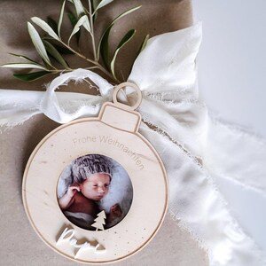 Pu&ograve; includere: Un ornamento natalizio in legno a forma di sfera con una cornice per foto al centro. L'ornamento ha il testo "Frohe Weihnachten" e "Papa" scritto su di esso. Un bambino &egrave; raffigurato nella cornice.