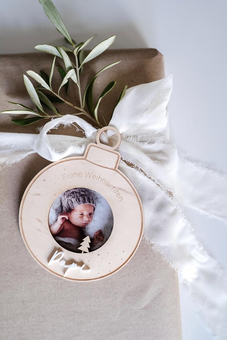Pu&ograve; includere: Un ornamento natalizio in legno a forma di sfera con una cornice per foto al centro. L'ornamento ha il testo "Frohe Weihnachten" e "Papa" scritto su di esso. Un bambino &egrave; raffigurato nella cornice.