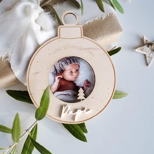 Pu&ograve; includere: Un ornamento natalizio in legno a forma di sfera con una cornice per foto al centro. L'ornamento &egrave; decorato con il testo "Frohe Weihnachten" e la parola "Pap&agrave;". Un bambino &egrave; ritratto nella cornice.
