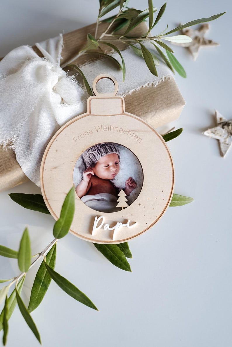 Pu&ograve; includere: Un ornamento natalizio in legno a forma di sfera con una cornice per foto al centro. L'ornamento &egrave; decorato con il testo "Frohe Weihnachten" e la parola "Pap&agrave;". Un bambino &egrave; ritratto nella cornice.