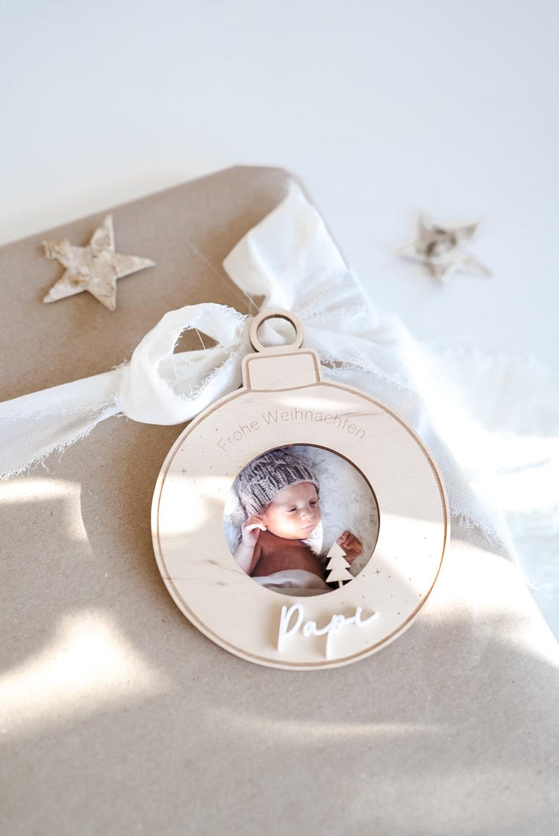 Pu&ograve; includere: Un ornamento natalizio in legno con una cornice per foto che mostra un bambino con un berretto di lana. L'ornamento &egrave; decorato con un piccolo albero e il testo "Frohe Weihnachten" e "Papi".