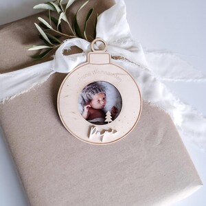 Pu&ograve; includere: Un ornamento natalizio in legno a forma di sfera con una cornice per foto al centro. L'ornamento ha il testo "Frohe Weihnachten" e "Papa" scritto su di esso. Un bambino &egrave; raffigurato nella cornice.