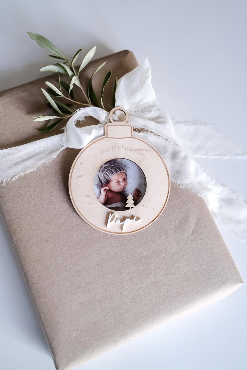 Pu&ograve; includere: Un ornamento natalizio in legno a forma di sfera con una cornice per foto al centro. L'ornamento ha il testo "Frohe Weihnachten" e "Papa" scritto su di esso. Un bambino &egrave; raffigurato nella cornice.