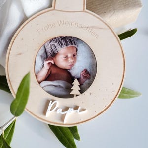 Pu&ograve; includere: Un ornamento natalizio in legno con una cornice per foto e il testo "Frohe Weihnachten" e "Dapi" in bianco. L'ornamento presenta una foto di un bambino con un berretto di lana.