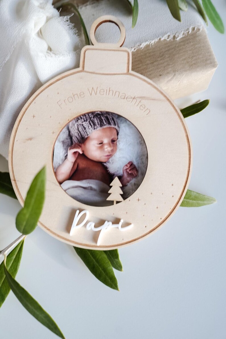 Pu&ograve; includere: Un ornamento natalizio in legno con una cornice per foto e il testo "Frohe Weihnachten" e "Dapi" in bianco. L'ornamento presenta una foto di un bambino con un berretto di lana.