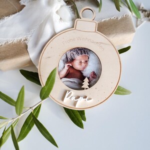 Pu&ograve; includere: Un ornamento natalizio in legno con una cornice per foto che mostra un bambino con un cappello. L'ornamento &egrave; decorato con stelle bianche e il testo "Frohe Weihnachten" e "Papi".