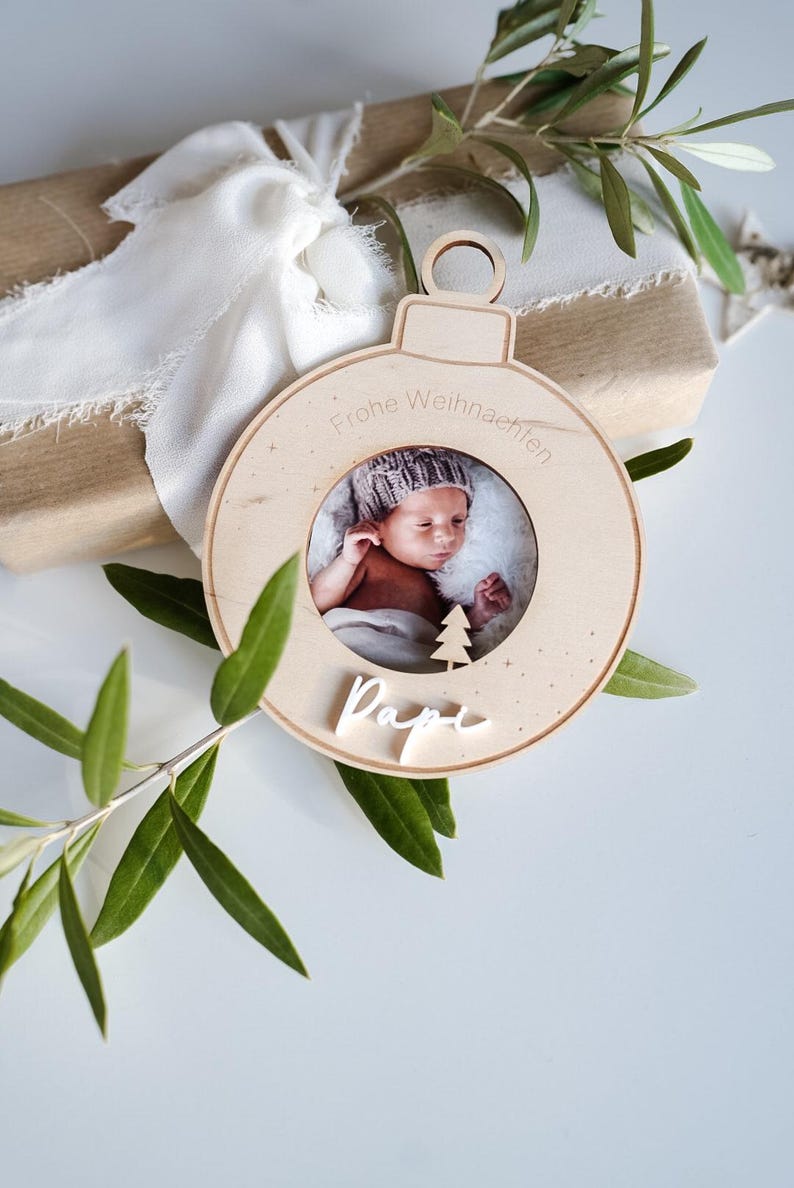 Pu&ograve; includere: Un ornamento natalizio in legno con una cornice per foto che mostra un bambino con un cappello. L'ornamento &egrave; decorato con stelle bianche e il testo "Frohe Weihnachten" e "Papi".