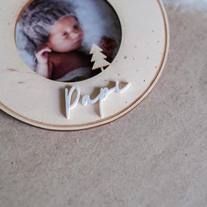 Pu&ograve; includere: Un ornamento natalizio in legno con una cornice per foto e la parola "Pappa" scritta in bianco. L'ornamento &egrave; decorato con un piccolo albero bianco e stelle.