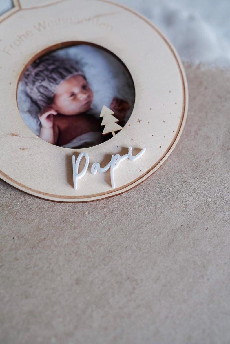 Pu&ograve; includere: Un ornamento natalizio in legno con una cornice per foto e la parola "Pappa" scritta in bianco. L'ornamento &egrave; decorato con un piccolo albero bianco e stelle.
