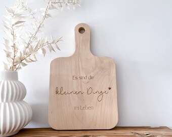 Può includere: Un tagliere di legno con il testo "Es sind die kleinen Dinge im Leben" (Sono le piccole cose della vita) inciso su di esso. Il tagliere ha un manico ed è appoggiato su uno scaffale di legno.