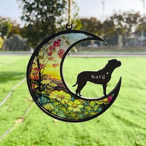 May include: A wooden crescent moon ornament with a stained glass design featuring a sunset, flowers, and a silhouette of a dog. The dog's name, "Sarg", is engraved on the ornament.