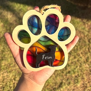 May include: A paw print-shaped wooden ornament featuring a multi-coloured stained glass-style insert. A black silhouette of a dog is layered over the centre, with the name Fern printed in white script. A jute twine loop is attached to the top.