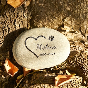 May include: A light gray, oval-shaped memorial stone with a heart and paw print design. The name "Melina" and the years "2005-2025" are inscribed on the stone. The stone is resting on a textured, brown surface with fallen leaves.