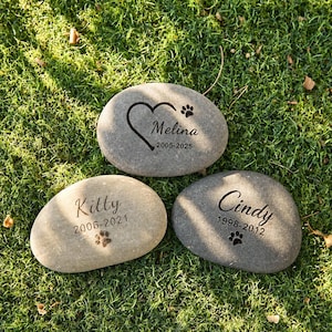 May include: Three smooth, oval-shaped grey memorial stones engraved with names and dates. One stone features a heart and paw print with the name Melina 2005-2025. The others are inscribed with Kitty 2006-2021 and Cindy 1998-2012, each including a paw print.