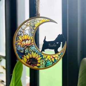 May include: A wooden crescent moon ornament with a sunflower design and a black silhouette of a dog with the name "Nala" engraved on it.