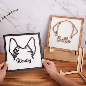 Può includere: Due cartelli in legno con ritratti di cani. Uno ha una cornice nera con il nome "Stouty" e una silhouette di cane. L'altro ha una cornice in legno con il nome "Bella" e una silhouette di cane. Entrambi sono su supporti in legno.