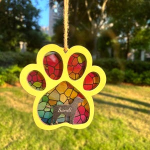 May include: A yellow paw print ornament with a stained glass effect. The ornament features a dog silhouette and the name "Sandi". It is suspended by twine. The background shows green grass and trees, creating a natural setting.