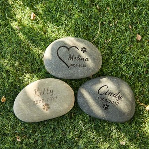 May include: Three engraved memorial stones for pets, each with a name, dates, and paw print. One stone has a heart design. The stones are gray and sit on green grass. The names are Melina, Kitty, and Cindy.
