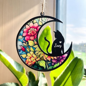 Puede incluir: Un adorno colgante en forma de media luna con marco negro. Un lado presenta un patrón floral estilo vidriera en rosa, amarillo, azul y morado. Una silueta de gato negro se sienta en la luna con el texto blanco Joey y un pequeño corazón.