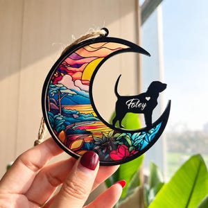 May include: A crescent moon-shaped ornament with a black frame and a colorful stained-glass style insert featuring a landscape of trees and flowers. A black silhouette of a dog stands on the moon with the name Foley and a small heart printed.