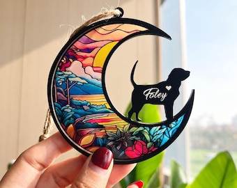 May include: A crescent moon-shaped ornament with a black frame and a colorful stained-glass style insert featuring a landscape of trees and flowers. A black silhouette of a dog stands on the moon with the name Foley and a small heart printed.