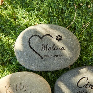May include: A gray stone memorial with the name "Melina", a heart, and a paw print engraved on it. The dates "2005-2025" are also engraved. The stone is set on a bed of green moss.