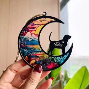 May include: A wooden crescent moon ornament with a stained glass design featuring a sunset over a mountain landscape. A black silhouette of a dog is inside the crescent moon with the name "Foley" engraved on it.
