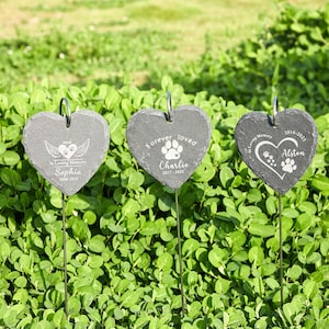 May include: Three heart-shaped slate garden markers on metal stakes. Each marker is inscribed with a different message: "In Loving Memory Sophia 2006-2025", "Forever loved Charlie 2017-2025", and "In Loving Memory Alston 2016-2023". The markers are set in green foliage.