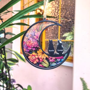Puede incluir: Un adorno decorativo de media luna con un diseño floral y dos siluetas de gatos. El adorno presenta los nombres "Sooty" y "Longy". El fondo está lleno de flores coloridas en tonos de rosa, amarillo y morado.