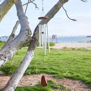 May include: A wind chime hangs from a tree branch. The chime has silver tubes, a wooden top and a wooden clapper. The background shows a grassy area, the ocean, and a bridge.