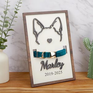 Puede incluir: Una placa de madera con una silueta de cabeza de perro, un collar de terciopelo azul verdoso con una hebilla dorada y el nombre "Marley" con las fechas "2019-2025" impresas debajo.