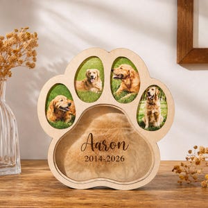 Può includere: Una cornice commemorativa in legno chiaro a forma di zampa con tre ritagli ovali per foto di golden retriever e un ampio scomparto centrale contenente pelo, con il testo stampato Aaron e le date 2014-2026.