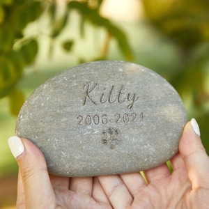 May include: A gray, oval memorial stone engraved with the name "Kitty", dates "2006-2021", and a paw print. The stone is held in a person's hands, with a blurred green background.
