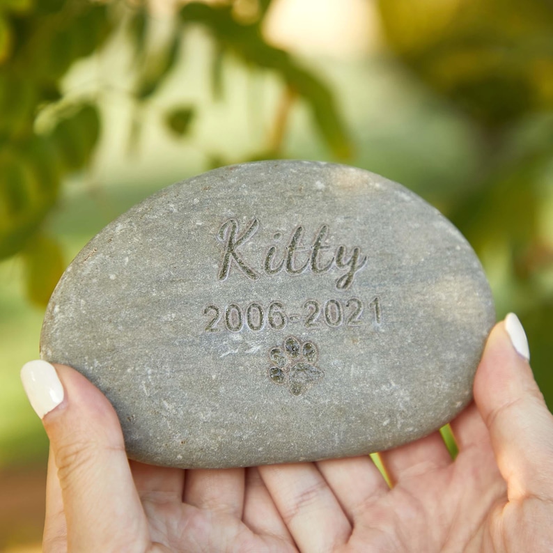 May include: A gray, oval memorial stone engraved with the name "Kitty", dates "2006-2021", and a paw print. The stone is held in a person's hands, with a blurred green background.