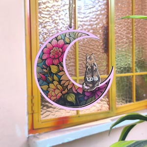 Könnte beinhalten: Ein rosa mondförmiger Ornament mit floralem Design und einer Katzen-Silhouette. Das Ornament hat einen farbenfrohen Buntglaseffekt mit rosa, gelben und lila Blumen. Die Katze ist braun und schwarz, und der Name "Lola" ist darauf geschrieben. Zum Aufhängen ist eine Juteschnur befestigt.