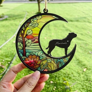 May include: A black metal crescent moon ornament with a stained glass design featuring a sunset and floral patterns. A black silhouette of a dog with the name "Marshall" is inside the moon.