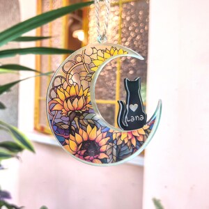 May include: A decorative crescent moon ornament featuring a black cat silhouette with a heart and the name "Lana". The moon is adorned with a stained-glass design of sunflowers in yellow, orange, and green.