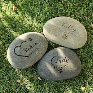 May include: Three engraved memorial stones for pets. Each gray stone features a pet's name and years of life. One stone has a heart and paw print, another has a paw print, and the last has a paw print.
