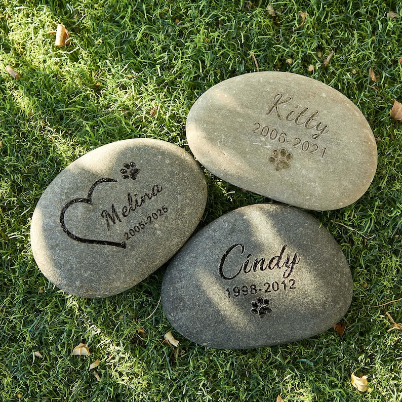May include: Three engraved memorial stones for pets. Each gray stone features a pet's name and years of life. One stone has a heart and paw print, another has a paw print, and the last has a paw print.