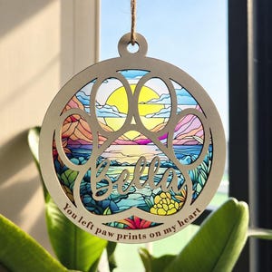May include: A circular wooden hanging ornament featuring a cutout paw print design. The centre displays a colourful stained-glass style landscape with a sun, mountains, and water. The name Bella and the text You left paw prints on my heart.