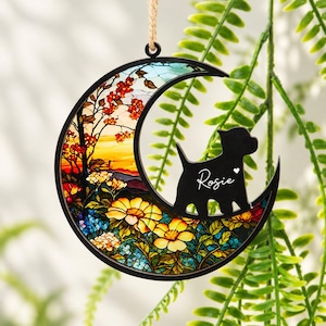 May include: A crescent moon-shaped hanging ornament with a black frame. The interior features a colourful stained-glass style landscape with yellow flowers, red blossoms, and a black silhouette of a dog with the name Rosie printed on it.